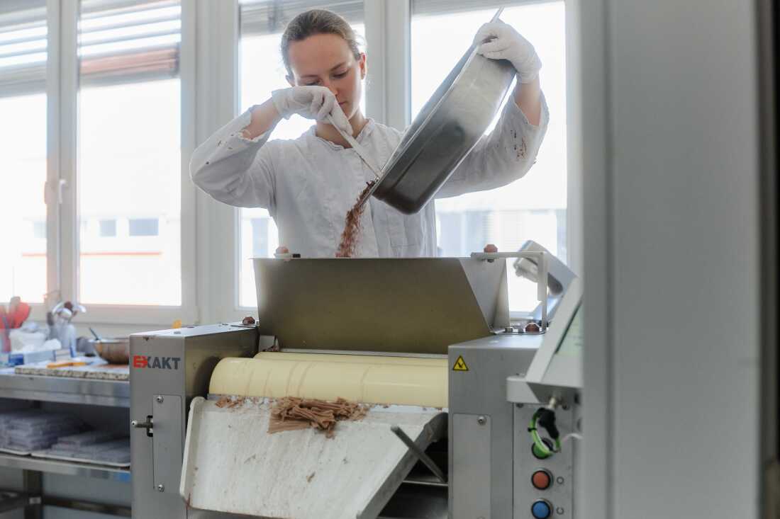 This photo shows an employee at the Planet A Foods testing lab testing and processing ChoViva. Dressed in a white top and white gloves, she holds a metal pan full of brown ChoViva seeds and uses a spatula to pour them into a funnel attached to a machine with two cylindrical rollers.