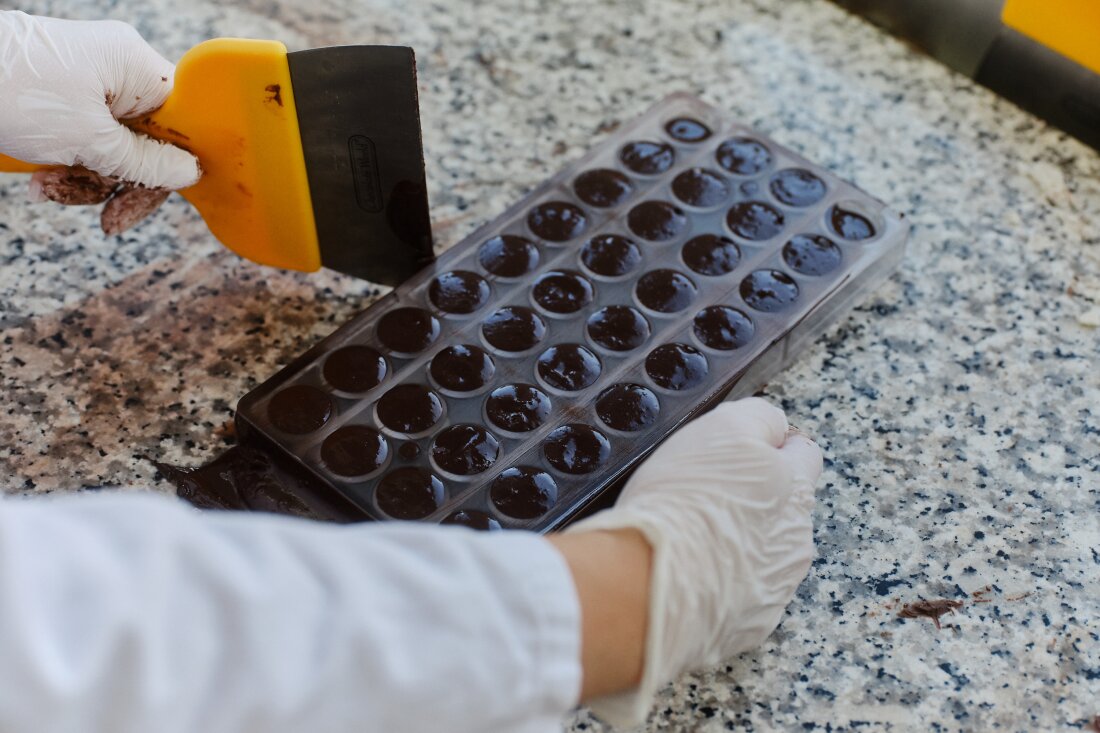 This photo shows the gloved hands of an employee in a Planet A Foods testing lab handling ChoViva, a cocoa-free chocolate alternative. The employee's right hand is on the right side of the tray, which has several rows of round molds that give ChoViva a sticky look. The employee's left hand holds a lyre.